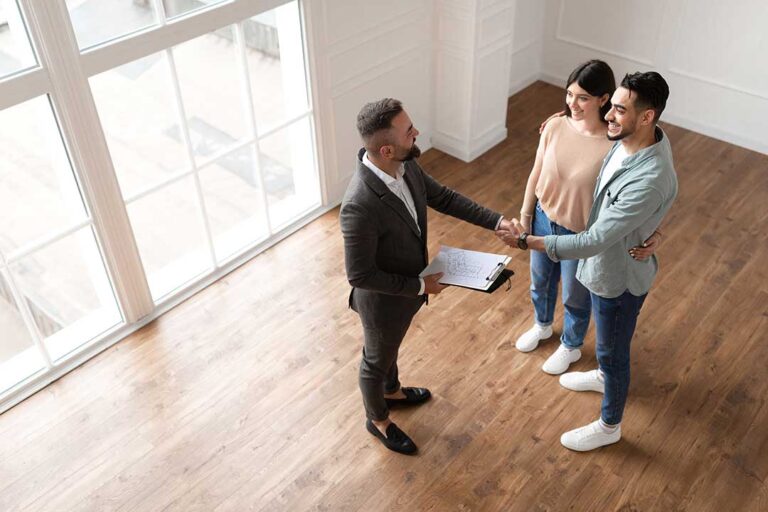 A buyer and real estate agent review property documents at a kitchen table with a calculator and notebook.